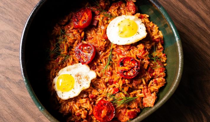 Fotografia em tons de marrom com um recipiente fundo e preto ao centro. Dentro do recipiente existe uma receita de arroz com linguiças. Em cima existe dois ovos de codorna fritos e tomates cereja tostados na frigideira.