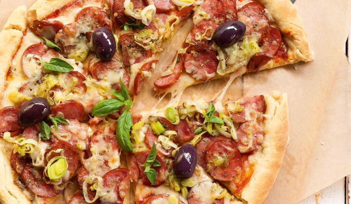 Fotografia em tons de marrom e vermelho de uma bancada de madeira branco com um paninho marrom claro, sobre ele uma pizza de calabresa e uma espátula retirando uma fatia. Ao lado um recipiente redondo amarelo com manjericão.