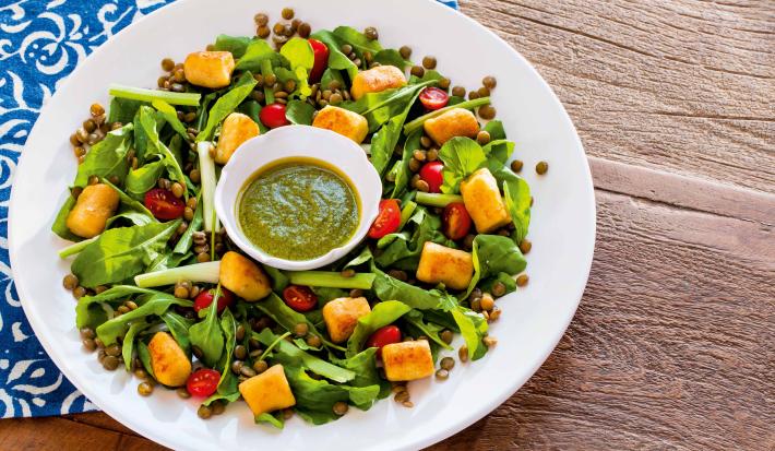 Fotografia em tons de verde em uma bancada de madeira com um pano azul desenhado, um prato branco raso redondo com a salada de cenoura, rúcula e tomate cereja.