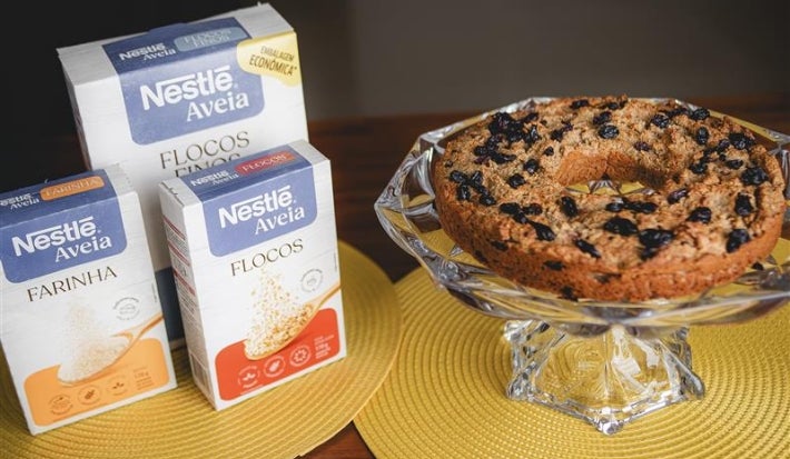 Fotografia em tons claros de três embalagens de aveia ao lado de um apoio de vidro com a receita do bolo inteiro.