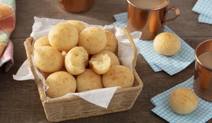 Fotografia em tons de azul em uma bancada de madeira escura, guardanapos de pano azul claro, uma cesta de vime com vários pães de queijo, xícaras cobre com café com leite dentro.