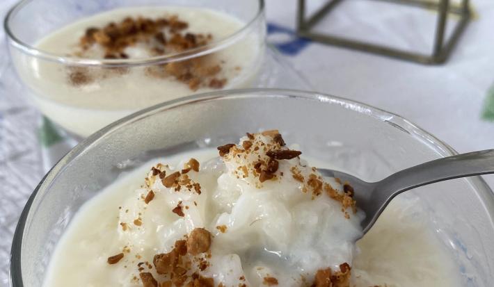 FotografiaFoto de um pote de vidro com a receita de Arroz Doce dentro, decorado com amendoins torrados. Há uma colher pegando um pouco do pote e ao fundo é possível ver a mesa decorada com uma toalha onde estão todos os itens da foto.