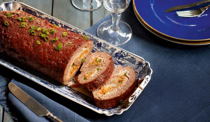 Fotografia em tons de azul escuro em uma mesa de madeira com uma travessa de prata ao centro com o bolo de carne recheado com vegetais Ao lado, um prato azul com garfo e faca apoiados nele e duas taças de vidro.