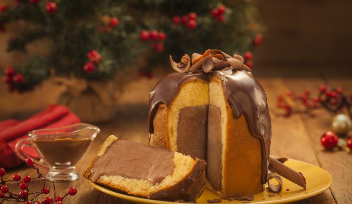Foto frontal de um panettone com uma fatia cortada. Ele está sobre um prato amarelo e dentro há um recheio de sorvete em tom marrom escuro. Na bancada de madeira há elementos de natal e um pequeno recipiente com calda de chocolate