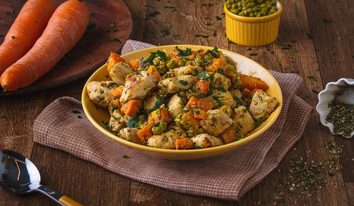 Imagem de uma bancada de madeira escura, sobre a qual há um prato amarelo com a receita de refogado de frango, outro prato com duas cenouras dentro e um pote pequeno com ervilhas verdes ao fundo