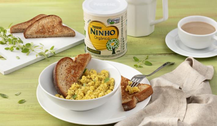 Fotografia em tons de verde e branco de uma bancada verde com um prato branco com um recipiente branco, sobre ele ovos mexidos e pão tostado. Ao lado um paninho beje, uma tábua com pão de forma tostado e orégano, uma lata de NINHO orgânico e uma xícara.