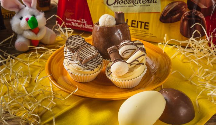 Foto aproximada de três miniovos de chocolate brancos, em um prato amarelo sobre uma bancada de madeira decorada com palha, um tecido amarelo, um pequeno coelho de pelúcia e uma barra de chocolate ao fundo