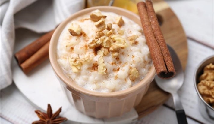 Em cima de uma mesa, recipiente redondo com arroz doce cremoso, com uma textura suave e aveludada, decorado com nozes picadas e canela em pau.
