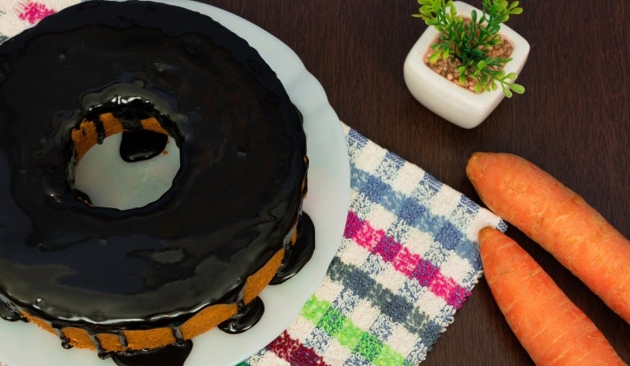 Bolo de cenoura com cobertura de chocolate, acompanhado de duas cenouras frescas e uma pequena planta decorativa. A composição é realçada por um pano xadrez colorido, tudo sobre uma superfície de madeira escura, transmitindo um ambiente acolhedor.