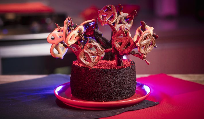 Bolo de chocolate decorado com galhos feitos de chocolate, formando uma árvore irregular onde ChocoPretzels ficam pendurados como pequenos frutos.