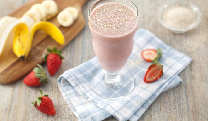 Fotografia em tons de cinza, azul, vermelho e amarelo de uma bancada de madeira cinza com um paninho xadrez azul e branco, sobre ele fatias de morango e um copo de vidro com a vitamina de banana e morango.