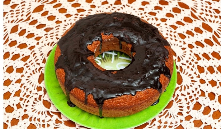 Bolo redondo recém-assado, com uma crosta dourada e cobertura de chocolate brilhante. Um pedaço foi cortado, revelando um interior amarelo úmido, e o bolo está sobre uma toalha de mesa de renda delicada.