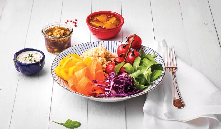 Fotografia em tons coloridos em uma bancada de madeira branca com um prato fundo com listras azuis e salada de rúcula, cenoura, cebola roxa e tomate dentro dele. Ao lado, potinhos com opções de molhos.