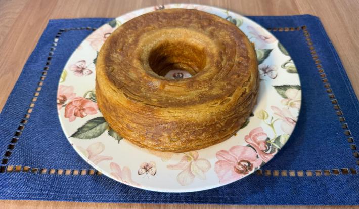 Fotografia de um bolo de café em um prato raso branco com desenhos de flores e folhas. O prato está sobre um pano azul quadrado apoiado em uma mesa de madeira.