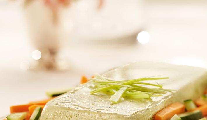 Fotografia em tons de branco, rosa e verde de uma bancada branca com uma bandeja rosa, sobre ela o terrine com palitos de cenoura e pepino. Ao fundo um vaso de flores rosa e uma jarra de água.