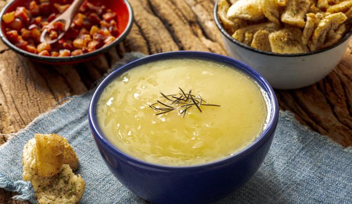 Fotografia de um recipiente fundo de vidro azul sobre um paninho azul claro apoiado em uma mesa de madeira. Dentro do recipiente tem caldo de aipim com um pouco de alecrim por cima, ao redor tem um pedaço de torrada caseira e amendoim.