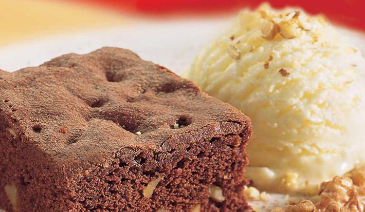 Fotografia em tons de marrom em uma bancada de madeira com uma foto aproximada de um prato polvilhado com cacau em pó, um brownie de chocolate e sorvete de creme.