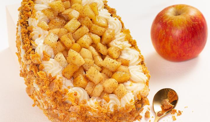 Foto da receita de Ovo Torta de Maçã, visto de cima, decorado com pedaços de maçã, massa de torta triturada, sobre uma bancada de mármore. Há uma maçã inteira e uma colher dourada na cena.