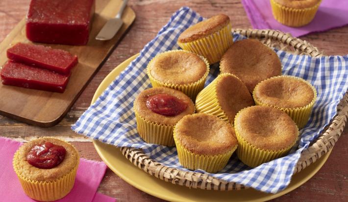 Fotografia em tons de marrom em uma bancada de madeira de cor marrom. No centro, um prato amarelo, uma cesta e um pano azul quadriculado em cima. Em cima do pano estão os cupcakes. Ao fundo, há uma tábua de madeira contendo fatias de goiabada e uma faca.