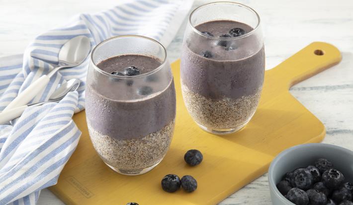 Receita de Pudim de Chia com Açaí e Mirtilos. Observa-se dois copos altos com uma parte de pudim de chia e outra parte de polpa de açaí, com mirtilos em cima. Os copos estão sobre uma tábua amarela e há um guardanapo de pano azul listrado sobre ela.