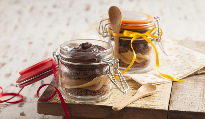 Fotografia em tons de marrom e cinza, com dois potes de vidro, um aberto e outro fechado contendo bolo fatiado, creme de chocolate e doce de leite sobre peça de madeira com colher e guardanapo ao lado, tudo sobre bancada em tons de cinza