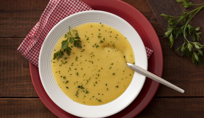 Fotografia em tons de branco, amarelo e vermelho, com prato branco ao centro contendo um creme amarelo com salsa e uma colher sobre guardanapo quadriculado em vermelho e branco e sous plat vermelho, ramo de salsa, e tudo sobre bancada de madeira rústica.