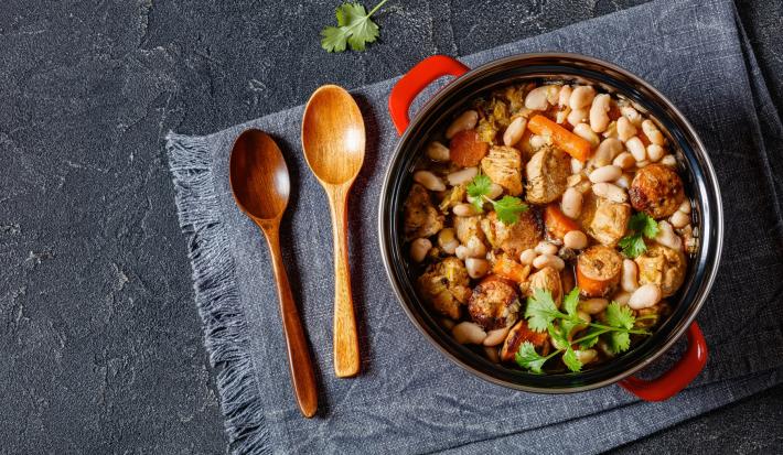 Imagem vista de cima de uma panela com cabo vermelho com a receita de cassoulet de feijão branco com linguiça e folhas de coentro decorando. Ao lado, duas colheres de pau e um pano azul escuro.