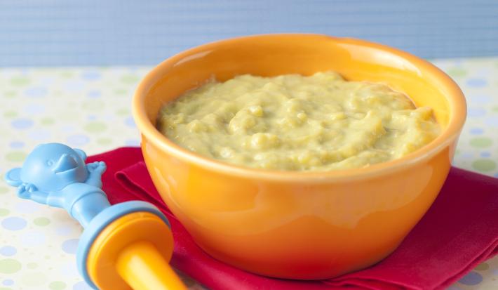 Fotografia em tons de amarelo, bordeaux e azul, com bowl amarelo contendo um creme verde claro sobre guardanapo bordeaux com uma colher infantil azul e amarela, tudo sobre toalha de bolinhas coloridas e fundo azul.