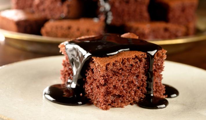 Imagem de um pedaço quadrado de bolo de chocolate sobre um prato claro com calda de chocolate escorrendo.