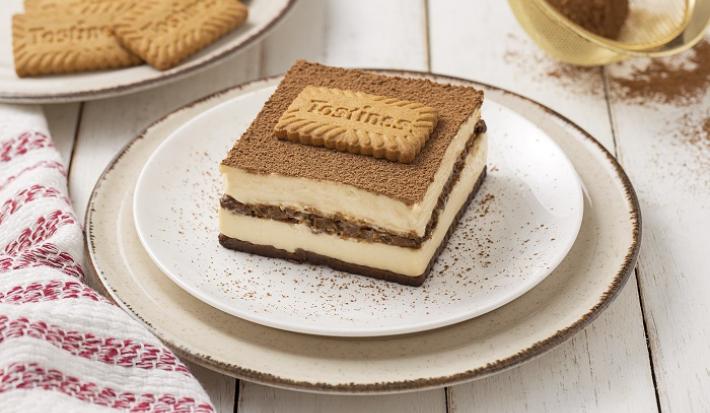 Foto da receita de Tiramisú de Tostines Especiarias. Observa-se uma fatia do Tiramisú bem alta expondo as camadas de creme e de biscoito. No topo, observa-se cacau em pó polvilhado com um Biscoito Tostines.