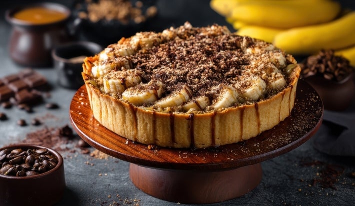 Em cima de uma mesa, banoffee com massa de biscoitos crocante, bananas em rodelas, polvilhado com canela em pó.