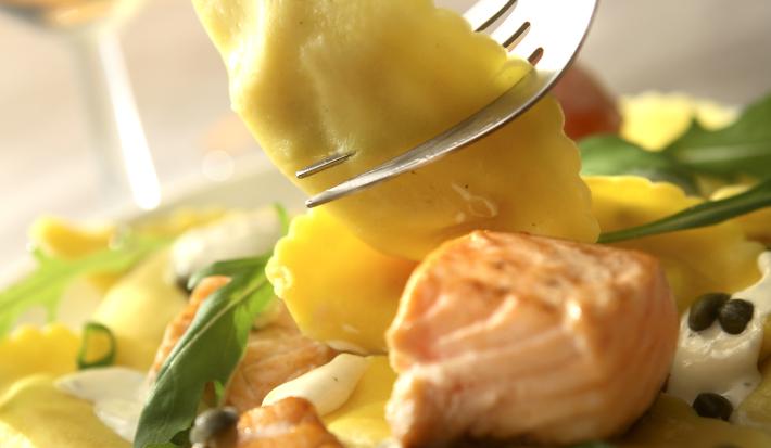 Fotografia de uma massa recheada com molho de creme de leite e salmão defumado, com grãos de pimenta e algumas folhas de rúcula dentro de um prato branco. A massa está sobre uma mesa cinza, e, ao fundo, duas taças com champanhe.