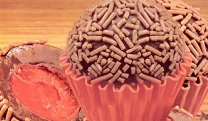 Fotografia em tons de marrom e vermelho de uma bancada de madeira com uma forminha vermelha, sobre ela brigadeiros recheados com cereja.