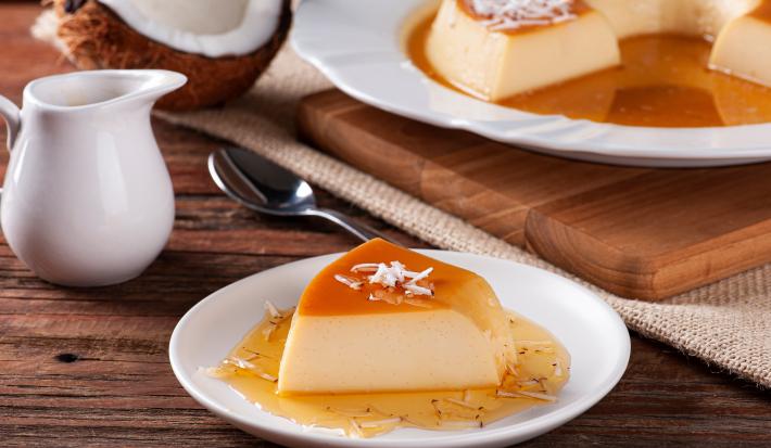 Fotografia de uma fatia de pudim de coco com caramelo salgado sobre um prato branco de vidro. Ao lado tem uma tábua de madeira com outro prato branco e o resto do pudim.