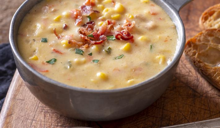 Foto aproximada de uma tigela cinza com a receita de sopa de milho em tom amarelo, sobre uma tábua de madeira ao lado de uma colher de metal.