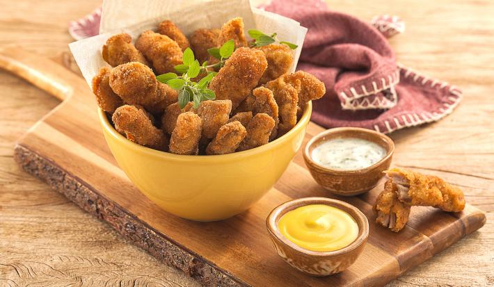 Fotografia em tons de amarelo em uma bancada de madeira clara, um paninho roxo, uma tábua de madeira com um potinho amarelo em cima e várias iscas empanadas de carne suína dentro dele. Ao lado, potinhos com mostarda e molho tártaro.