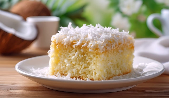 Fotografia de um bolo cortado em quadrado em um prato branco, com interior amarelo fofinho, úmido e cobertura de coco. O fundo natural com folhas verdes e um coco cortado.
