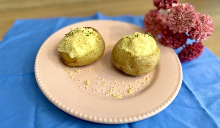 Fotografia em tons de azul e rosa com um prato rosa ao centro. Dentro do prato existe duas batatas recheadas com creme de ASCENDA e polvilhadas com queijo parmesão