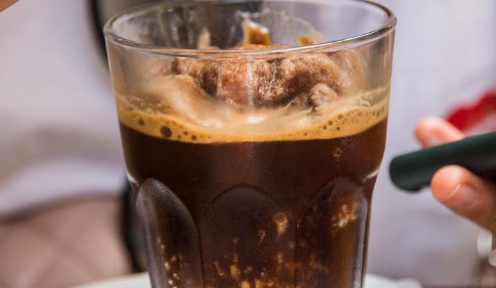 Fotografia de uma granita feita de café, leite e gelo que está em um copo de vidro, o qual está apoiado sobre um miniprato de vidro branco. Ao fundo, tem alguém segurando uma colher com a sobremesa.