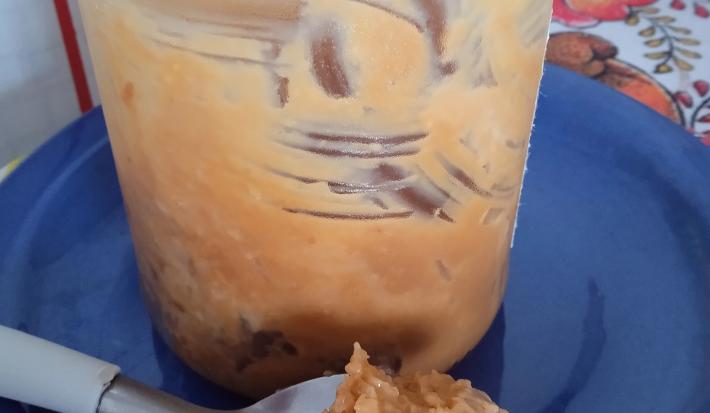 Fotografia de um pote de vidro com doce de leite caseiro com coco sobre um prato raso azul. Na frente do vidro tem uma colher com doce de leite e coco ralado.