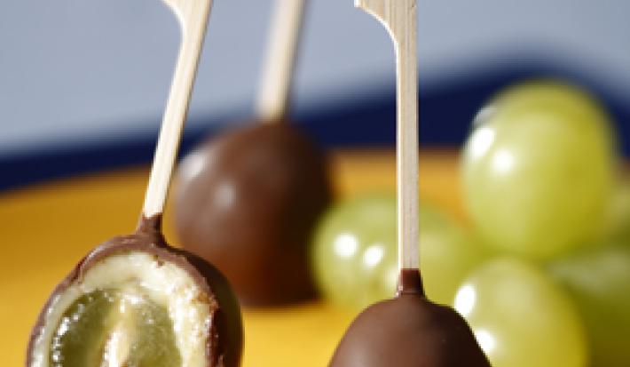 Fotografia em tons de amarelo, chocolate e azul, mostrando sobre bandeja amarela três espetinhos com chocolate, sendo um aberto e recheado com uva, algumas uvas verdes, tudo sobre guardanapo e bancada em tons de azul.