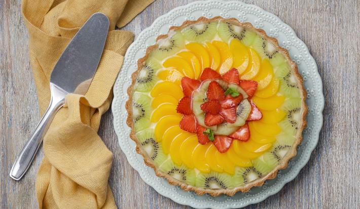 fotografia em tons de cinza, amarelo, verde e branco de uma bancada cinza vista de cima. Ao centro um prato redondo branco com uma torta com pedaços de frutas por cima para decorar, e ao lado um pano pardo cm uma espátula.