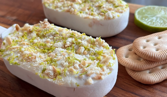 A imagem mostra dois sorvetes estilo picolé, claros, com uma textura crocante de biscoitos triturados e raspas de limão na superfície. Estão apoiados em uma tábua de madeira com 3 biscoitos de maisena e uma rodela de limão ao lado.