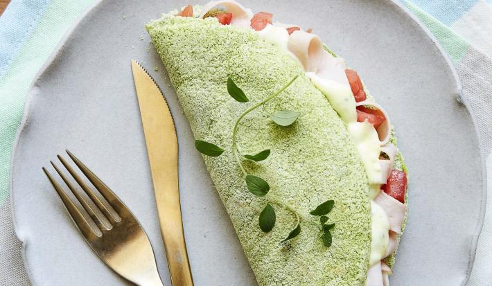 Fotografia em tons de verde em uma mesa de madeira com um pano listrado colorido em tons claro, um prato lilás redondo raso com uma tapioca verde de espinafre em cima dele.
