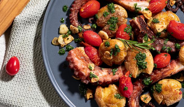 Imagem vista de cima de um prato em tom cinza com a receita de polvo, servido com batatas, tomates e algumas folhas verdes pequenas. O prato está sobre uma toalha clara e ao lado há um tábua de madeira e um tomate pequeno