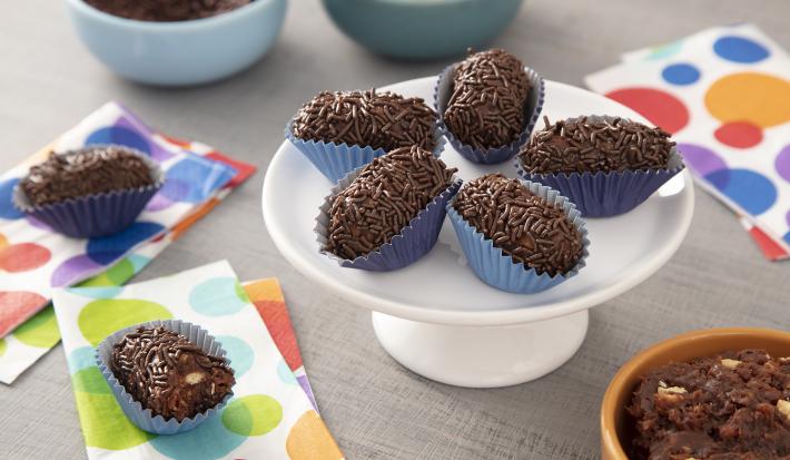 Foto da receita de Brigadeiro Croquete. Observa-se uma boleira pequena com 5 brigadeiros em formato de croquete em cima. A foto é decorada com guardanapos estampados de bolas coloridas e ao fundo observa-se dois potinhos: com brigadeiro e biscoito maria