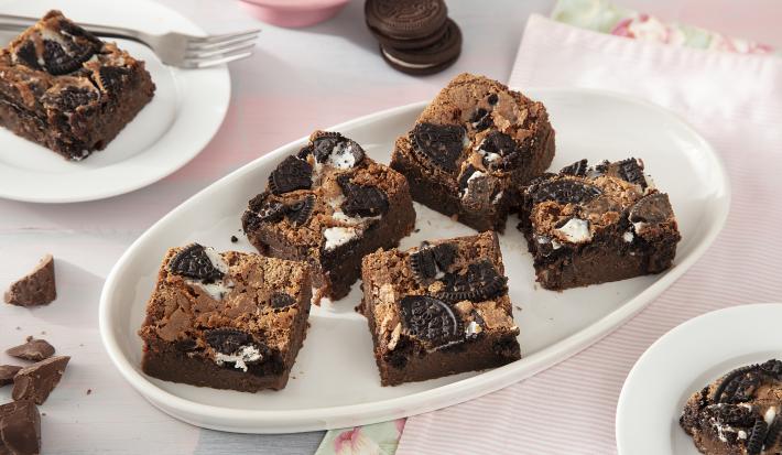 Foto da receita de Brownie de Leite Moça e Biscoito Negresco. Observa-se 7 fatias de brownie cortadas e dispostas em uma forma branca oval em um fundo rosa pastel.