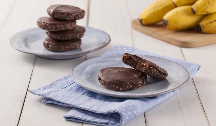 Fotografia em tons de marrom em uma bancada de madeira de cor branca. Ao centro, um pano azul claro estendido com um pires em cima contendo os cookies. Ao fundo, há mais um pires contendo cookies e uma tábua de madeira com algumas bananas.