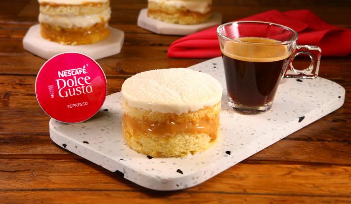 Fotografia em tons de vermelho em uma bancada de madeira escura, uma tábua de pedra branca com uma sobremesa de mini bolinho recheado com leite em pó e doce de leite. Ao lado, uma cápsula de Nescafé Dolce Gusto e uma xícara com o café pronto.
