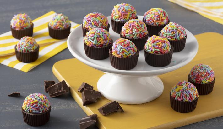 Fotografia em tons de amarelo em uma bancada cinza escura com guardanapos amarelos listrados. Ao centro, uma tábua amarela com um suporte para docinhos branco apoiando as forminhas de chocolate com brigadeiros com confeitos coloridos.